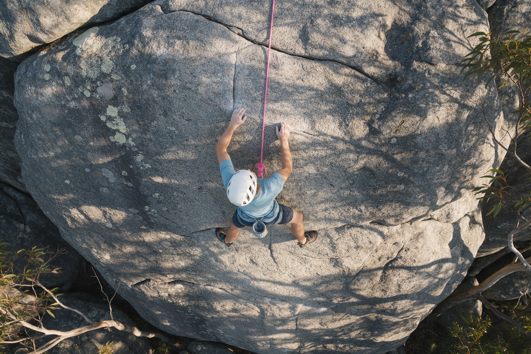 Looking straight down on a climber in a white helmet and blue shirt moving up a grey granite slab, with a pink rope running up from their harness.