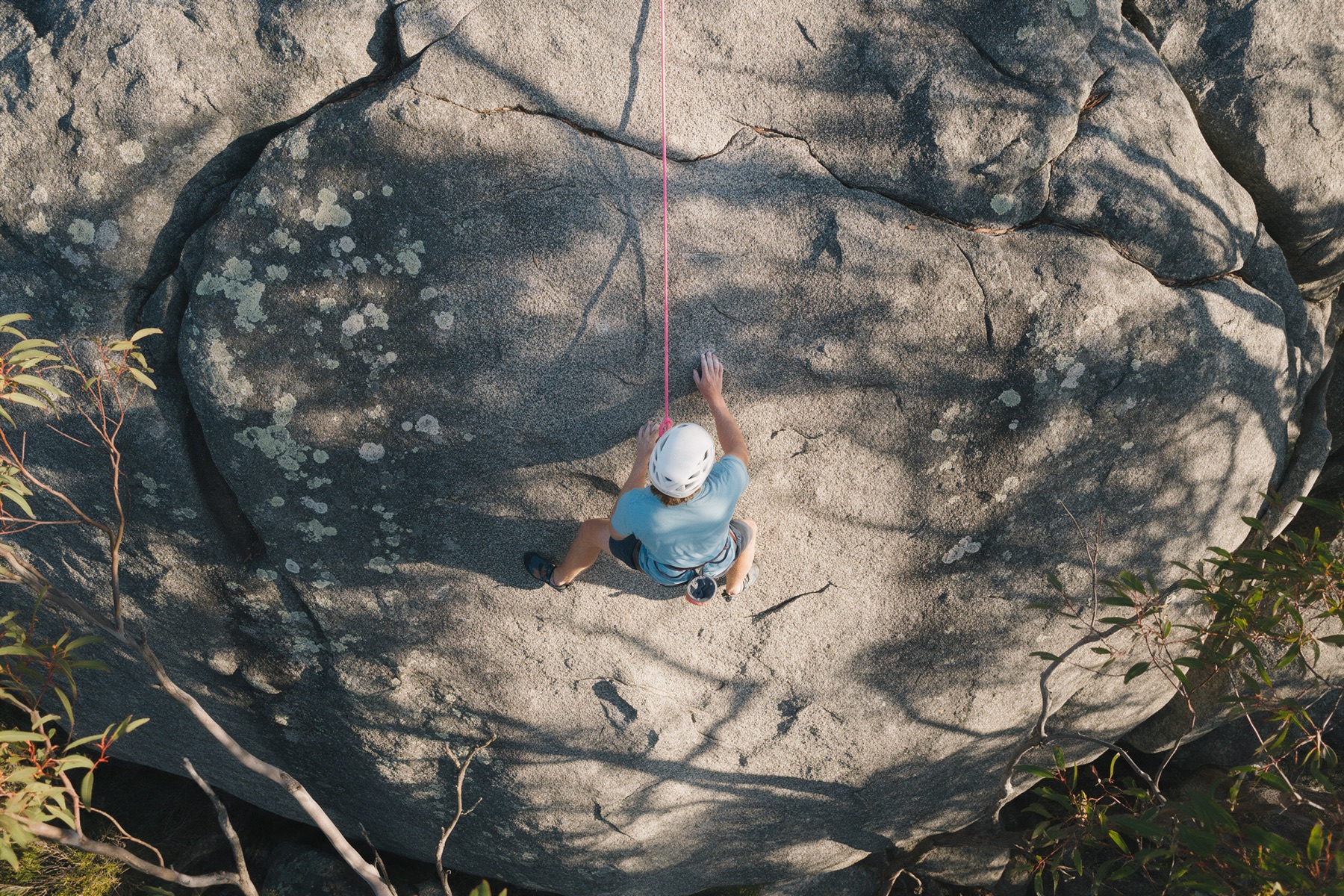 A climber on a wide grey granite slab surrounded by bush, seen from above.